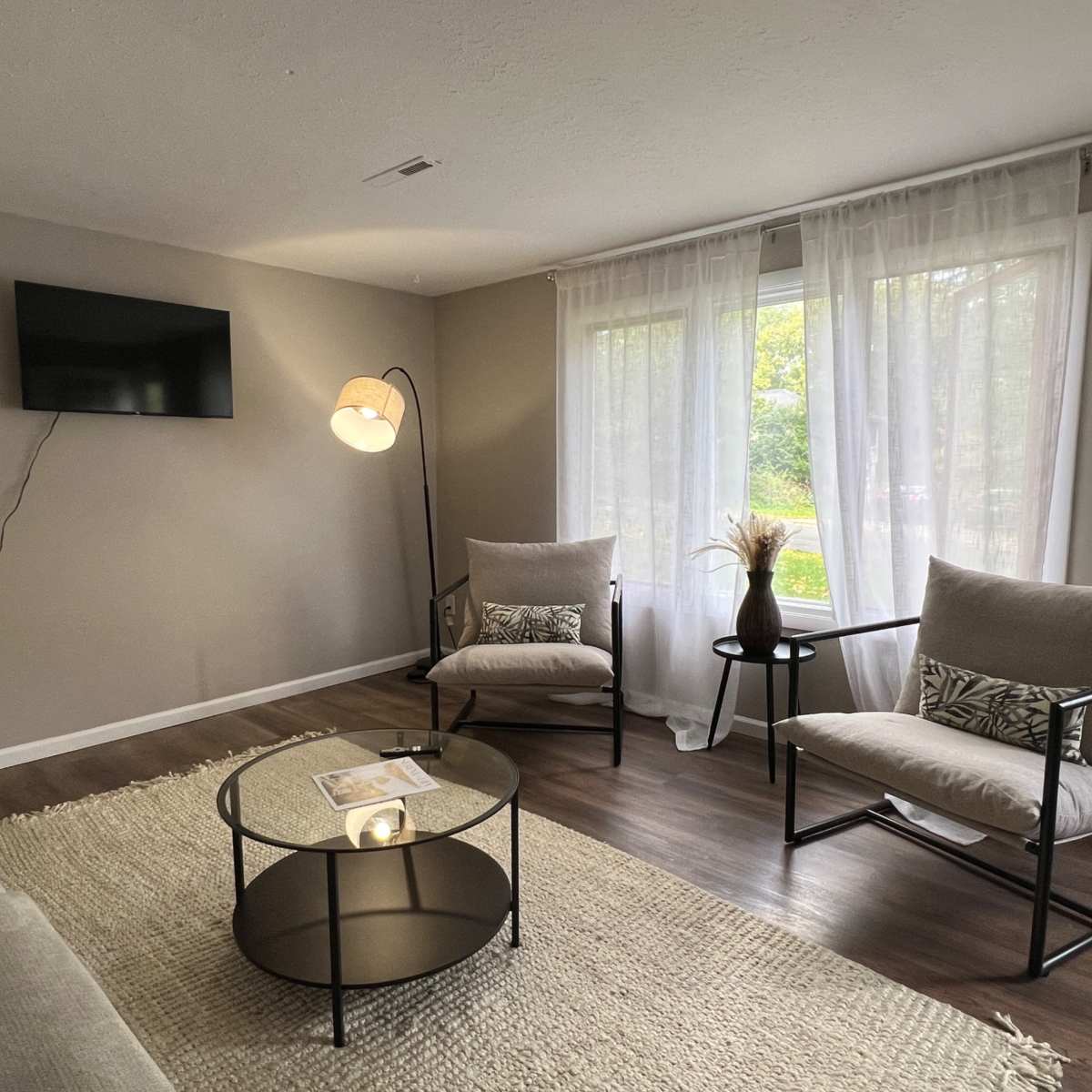 A cozy living room features a television mounted on the wall, two armchairs, a round coffee table, and large windows with sheer curtains allowing natural light.
