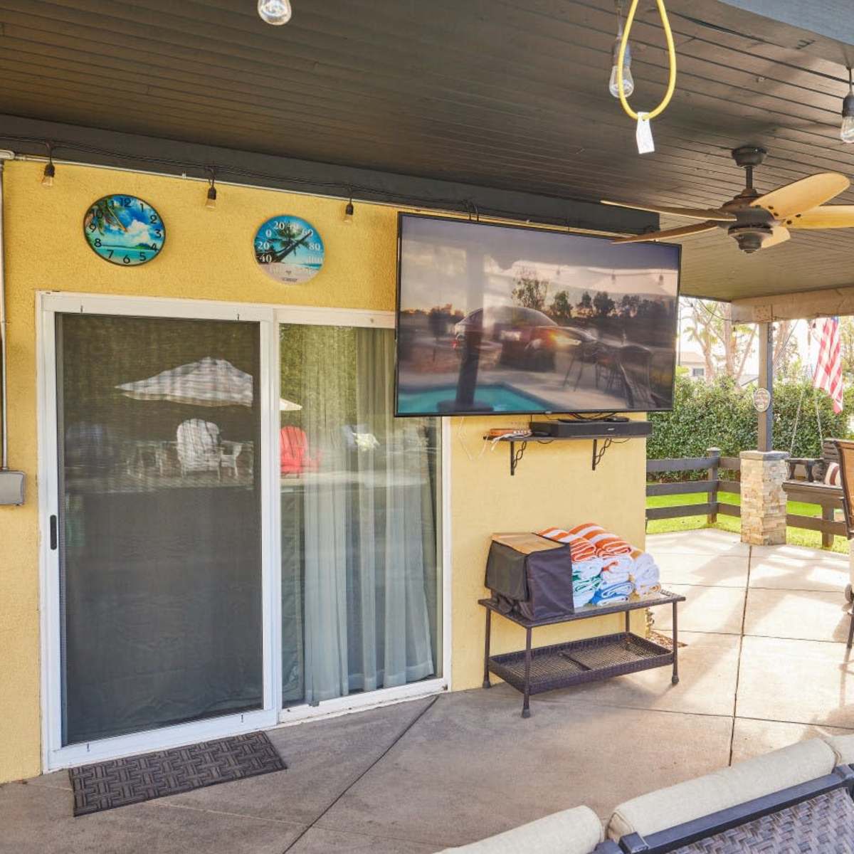 A patio area with a mounted TV, ceiling fan, and outdoor seating, surrounded by greenery and notable decor.
