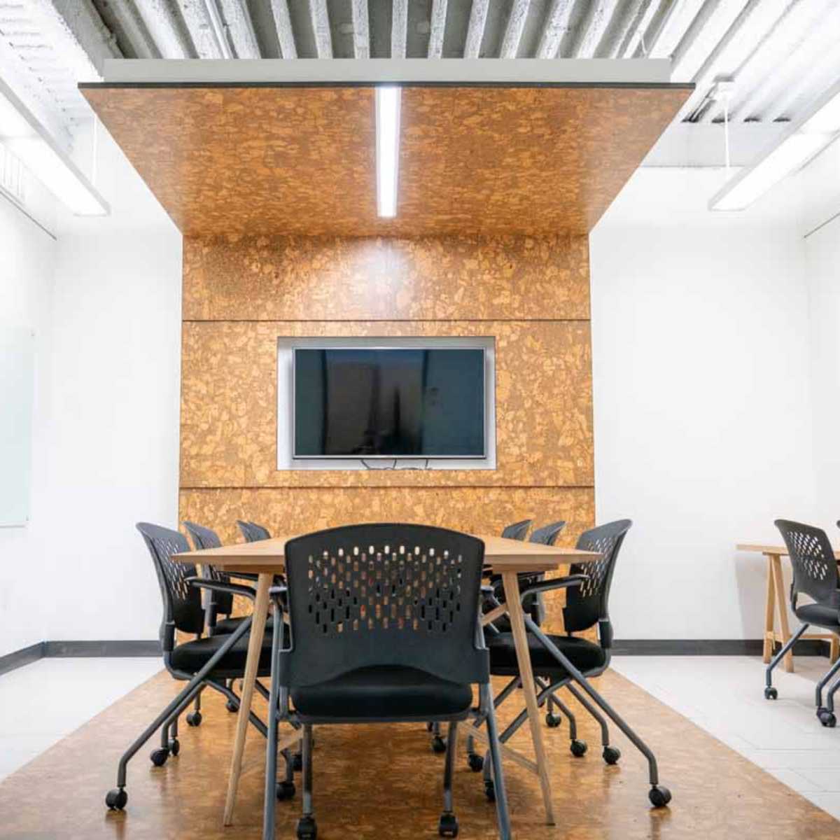 A modern conference room features a central table surrounded by black chairs, with a wall-mounted television and cork paneling in the background.