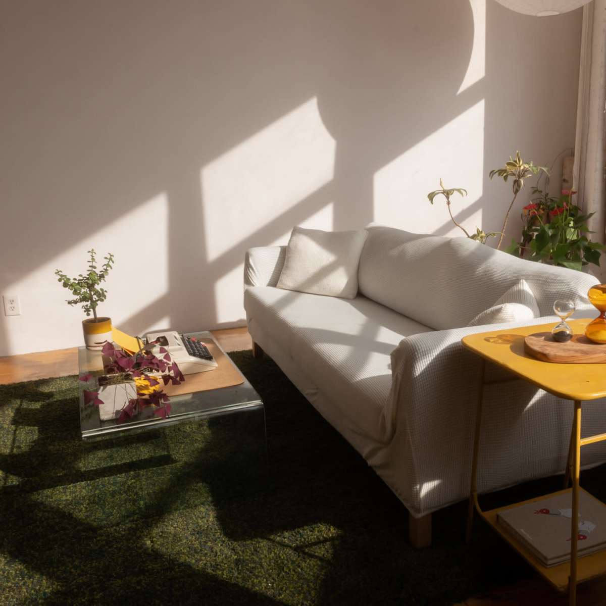 A white sofa is positioned in a sunlit room with plants and a yellow side table holding refreshments.