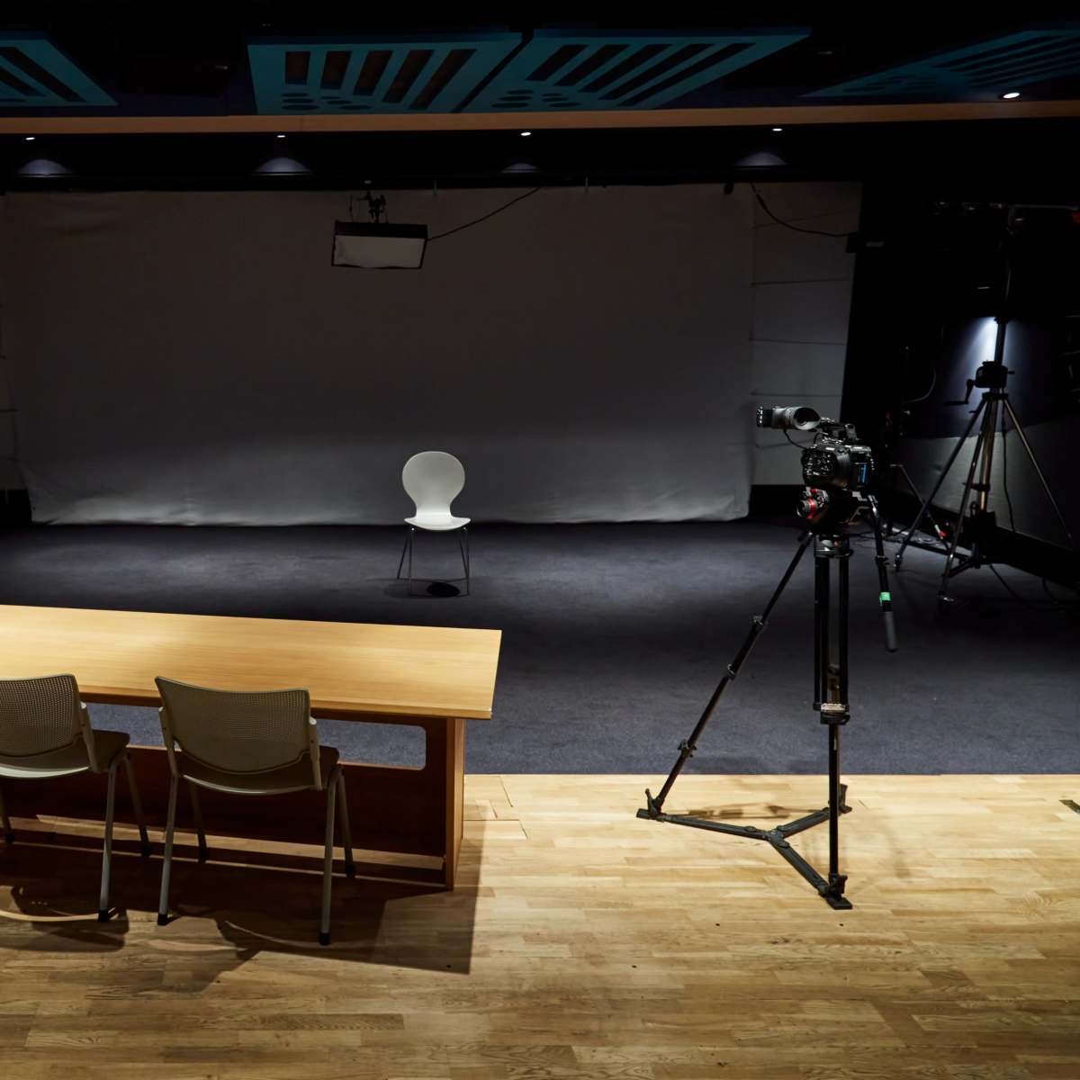 A dimly lit studio space with a wooden table and two chairs facing a spotlighted empty chair against a blank backdrop, surrounded by camera equipment.