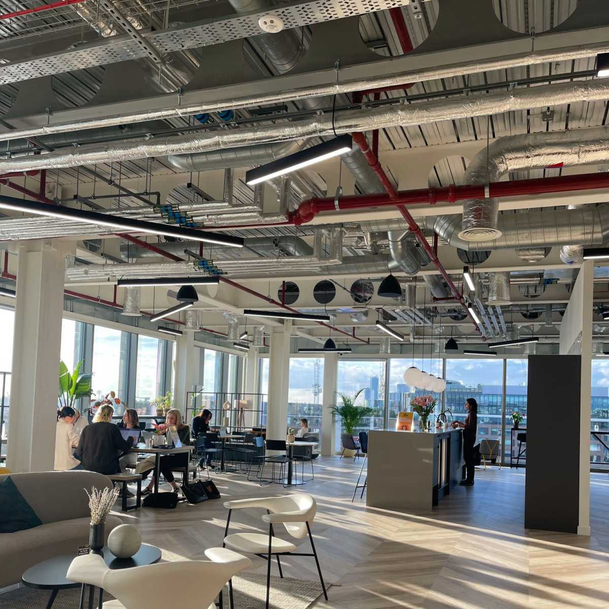 A modern office space with a seating area, a kitchen counter, and groups of people engaged in conversation near large windows.