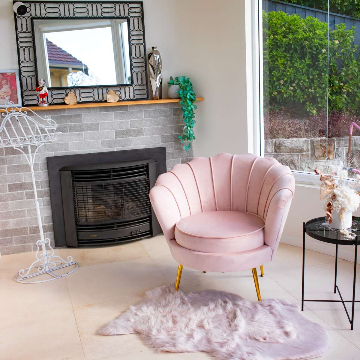 A pink velvet armchair is positioned next to a black glass side table in a bright corner of a room with a fireplace and large windows.