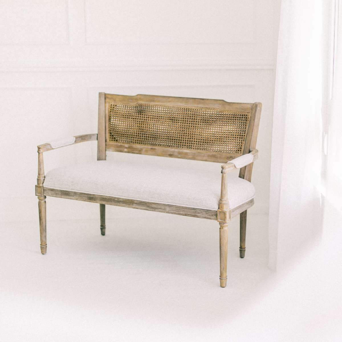 A wooden bench with a caned back and upholstered seat is positioned next to a window with sheer curtains.
