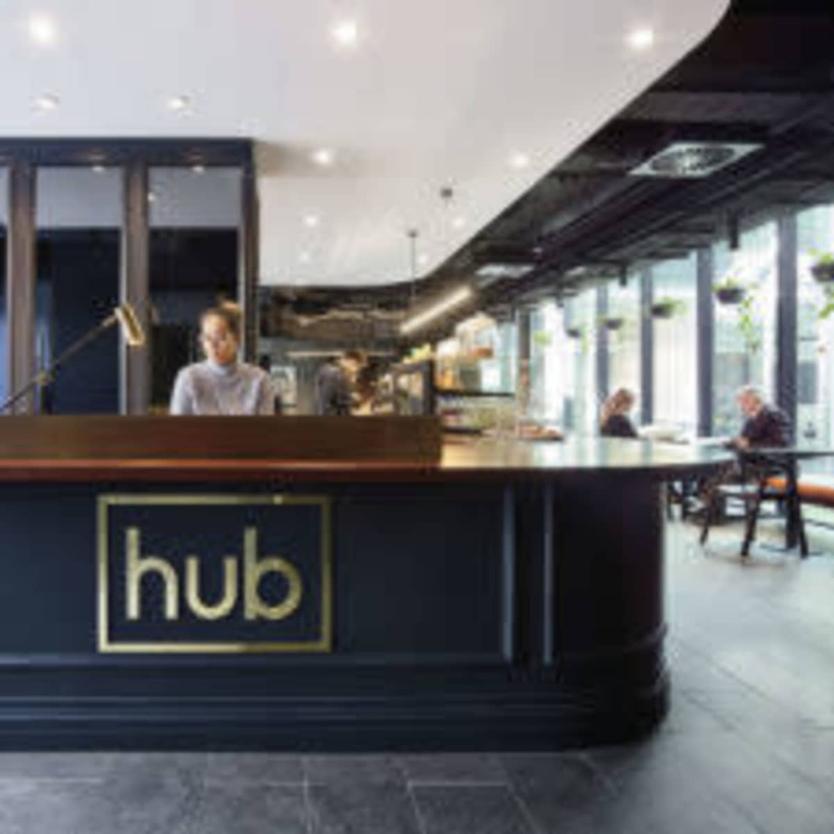 A sleek reception desk with the word "hub" prominently displayed, staffed by a person, in a modern, well-lit interior with seating areas in the background.