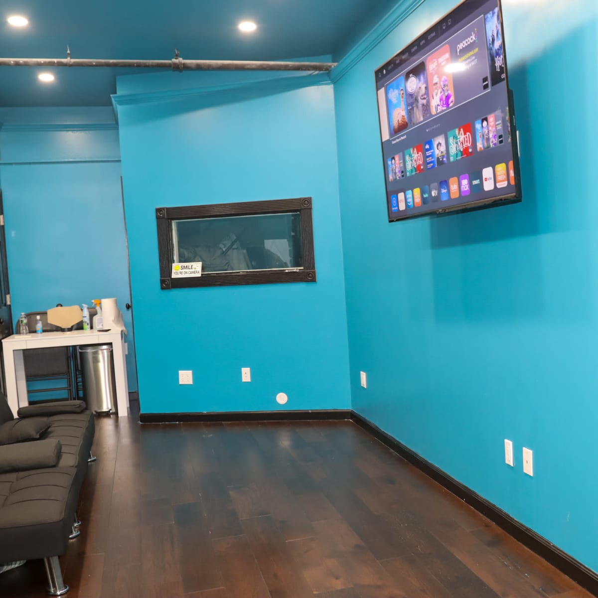 A bright blue room with a wall-mounted television displaying apps, a black sofa on the left, and an observation window on the right.