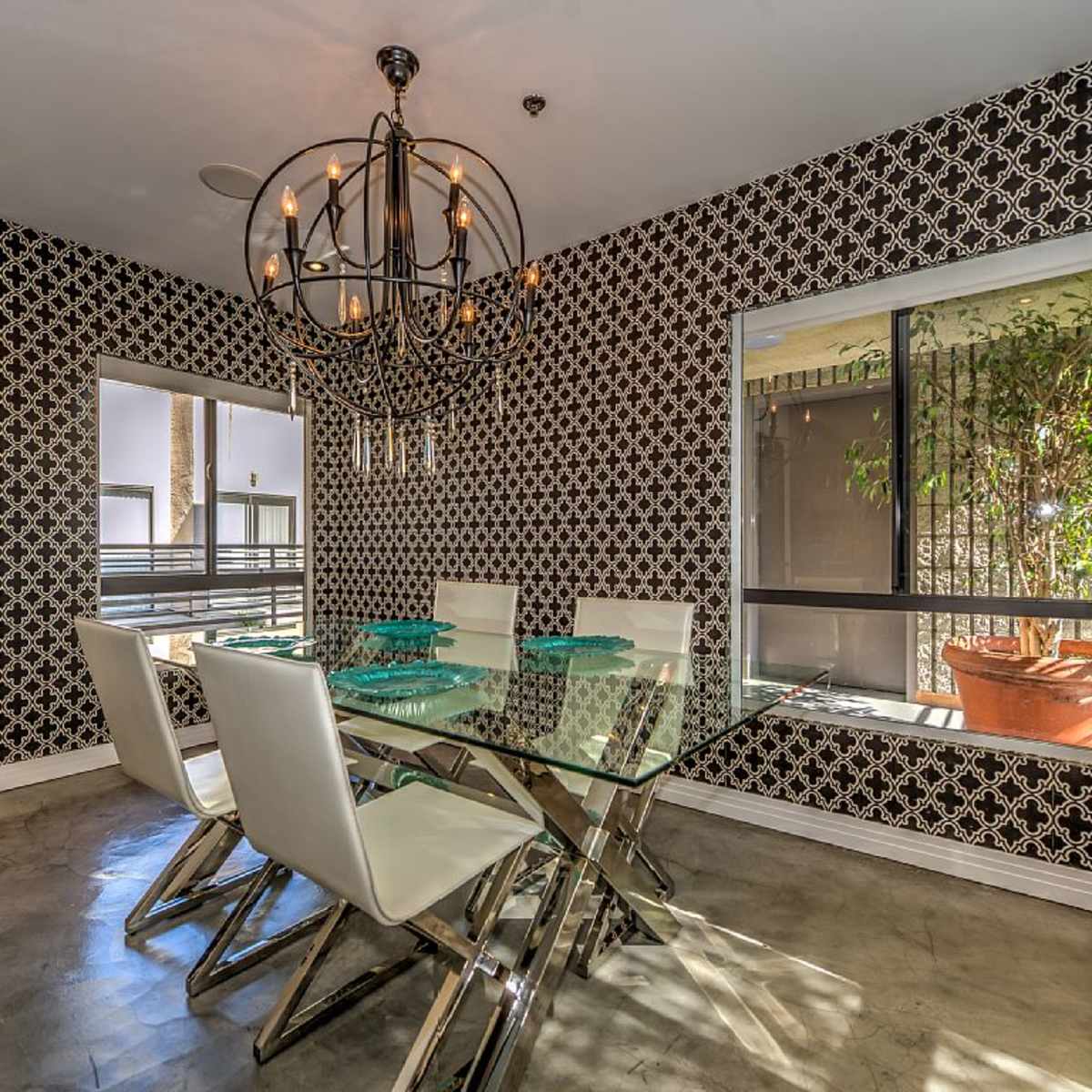 A dining area with a glass table surrounded by white chairs, set against a patterned black-and-white wall and featuring two windows with plants outside.