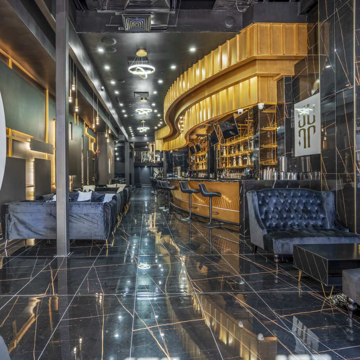 A modern bar interior with a polished black marble floor, elegant seating areas, and a long, illuminated bar counter in the background.