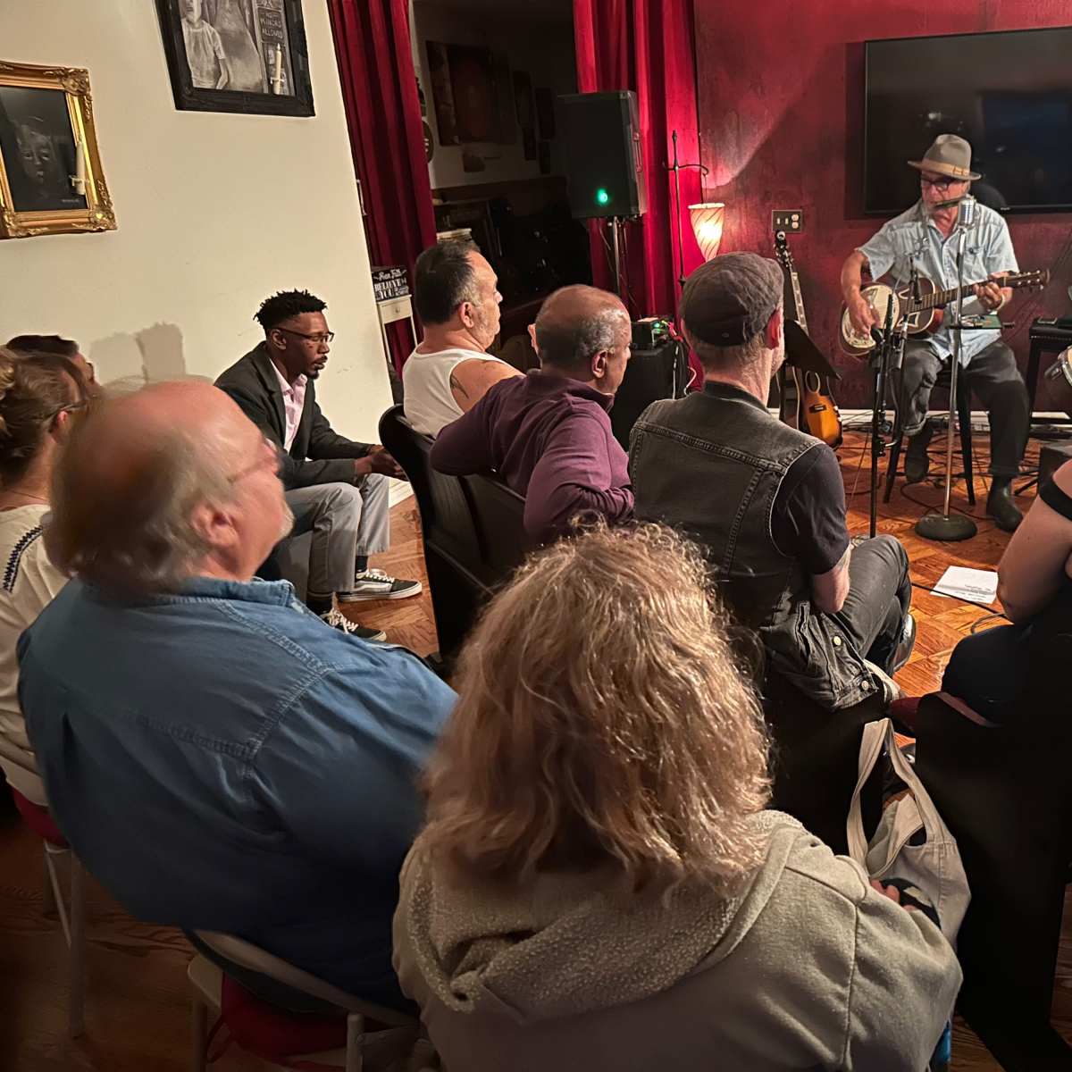 A musician plays guitar on stage while an audience of seated listeners watches attentively in a cozy room with red curtains.