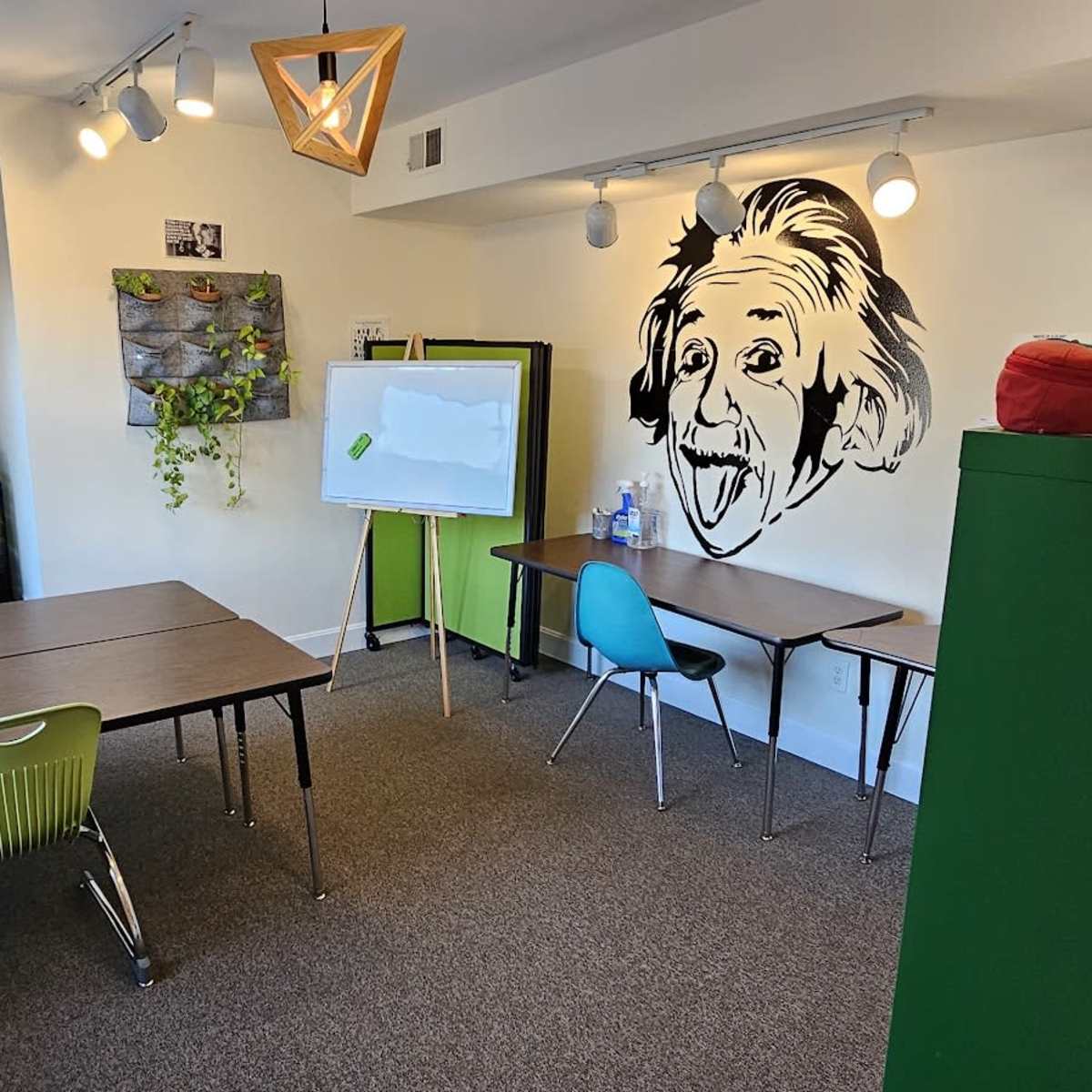 A classroom features several tables and chairs, a whiteboard, and a wall mural of Albert Einstein.