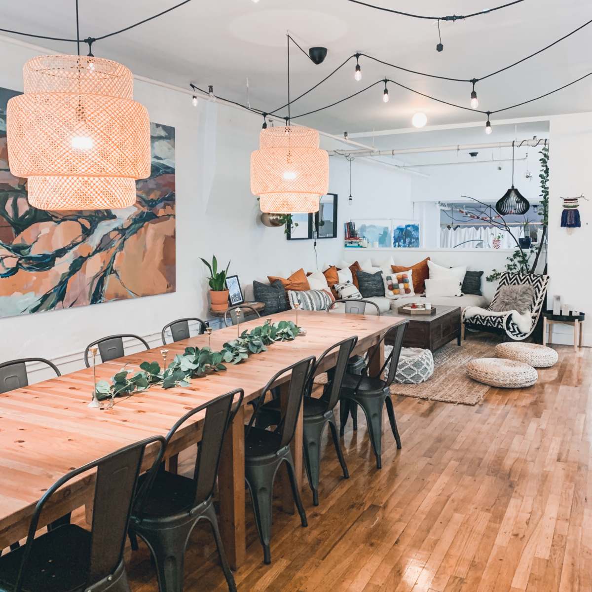 A spacious dining area featuring a long wooden table with metal chairs, pendant lights hanging overhead, and a cozy seating area with cushions and artwork on the walls.