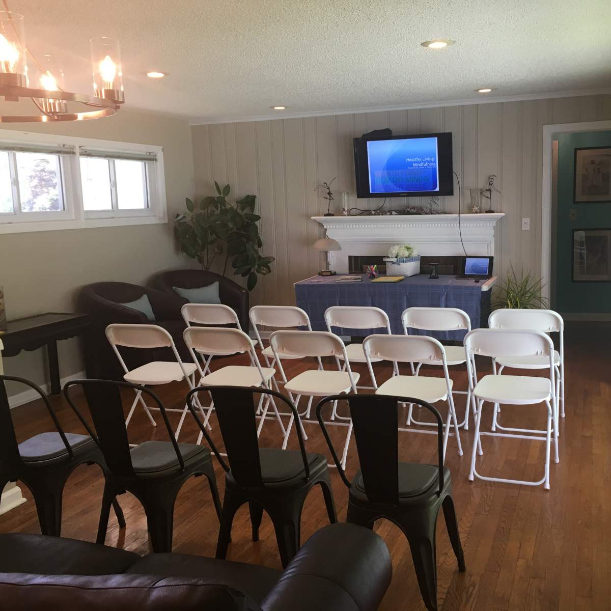 A living room is set up with rows of white folding chairs facing a table and a wall-mounted screen.