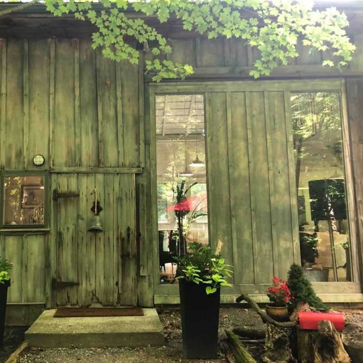 A wooden cabin with a green, weathered exterior, featuring a small door, a clock, and two planters on either side of the entrance.
