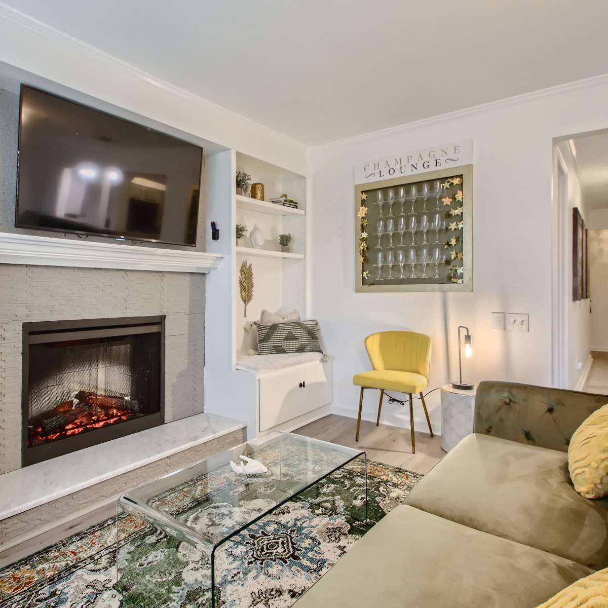 The living room features a modern fireplace beneath a wall-mounted TV, with a yellow chair, a glass coffee table, and decorative shelves.