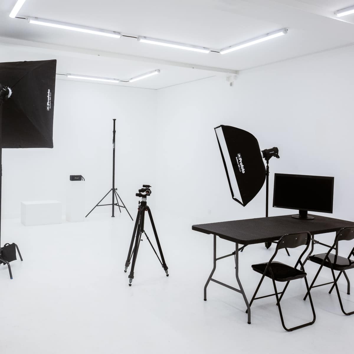 A photography studio with lighting equipment, a camera on a tripod, a monitor on a table, and two chairs in a minimalist white space.