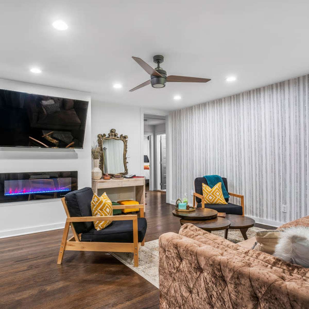 A modern living room features a wall-mounted TV, a sleek electric fireplace, and a mix of seating arrangements with decorative pillows.