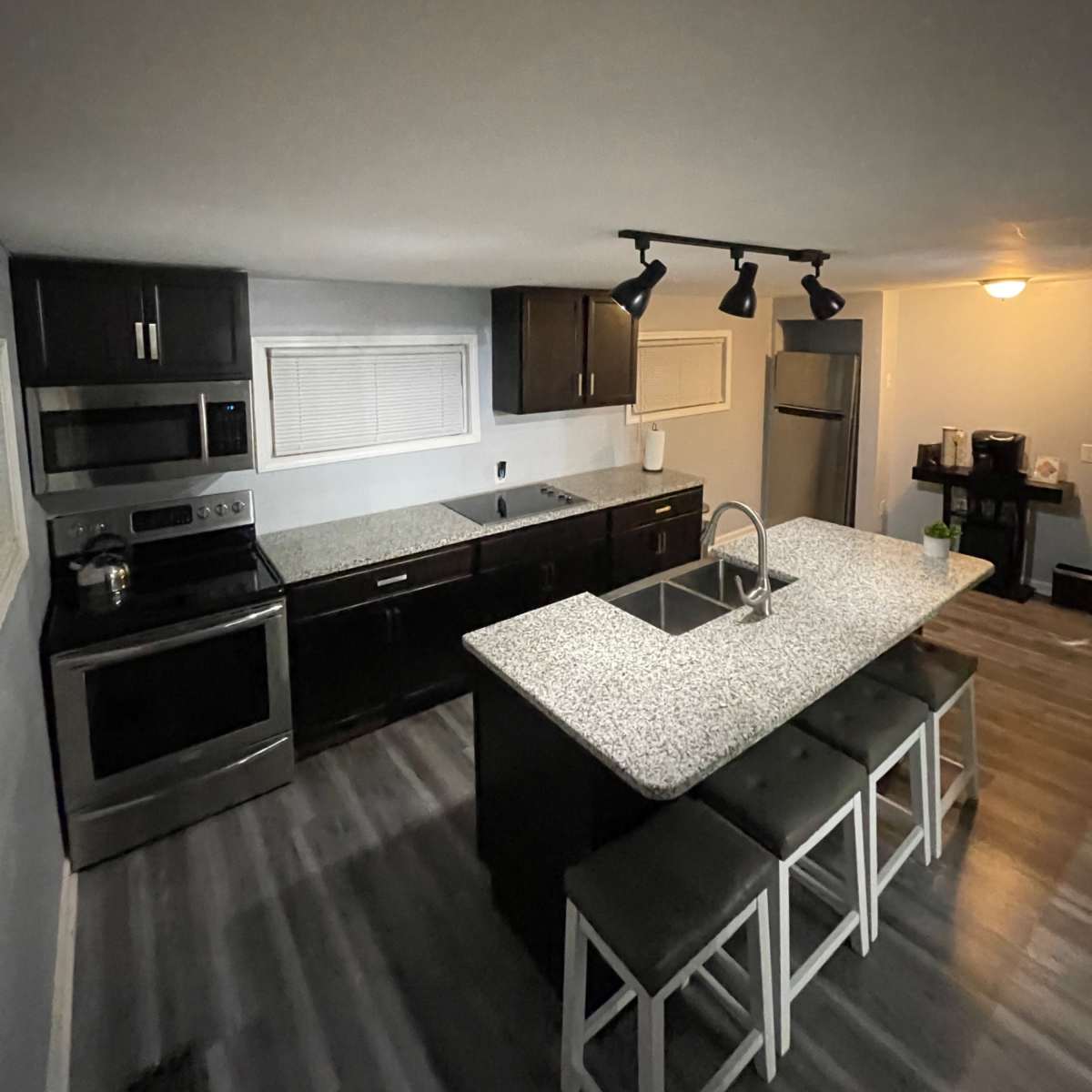 A modern kitchen features dark cabinetry, stainless steel appliances, a gray granite island with seating, and a light wood floor.