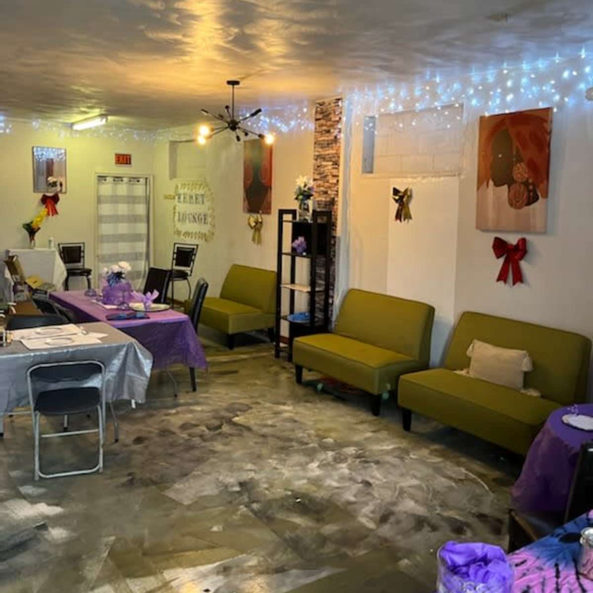 An interior of a small cafe or event space, with green couches, tables covered in purple and gray tablecloths, and decorative lights on the walls.