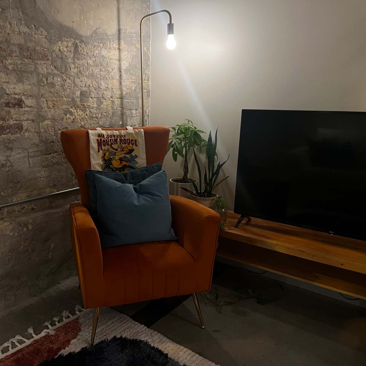 An orange armchair with a patterned throw sits beside a floor lamp and a television on a wooden stand, against a brick wall.