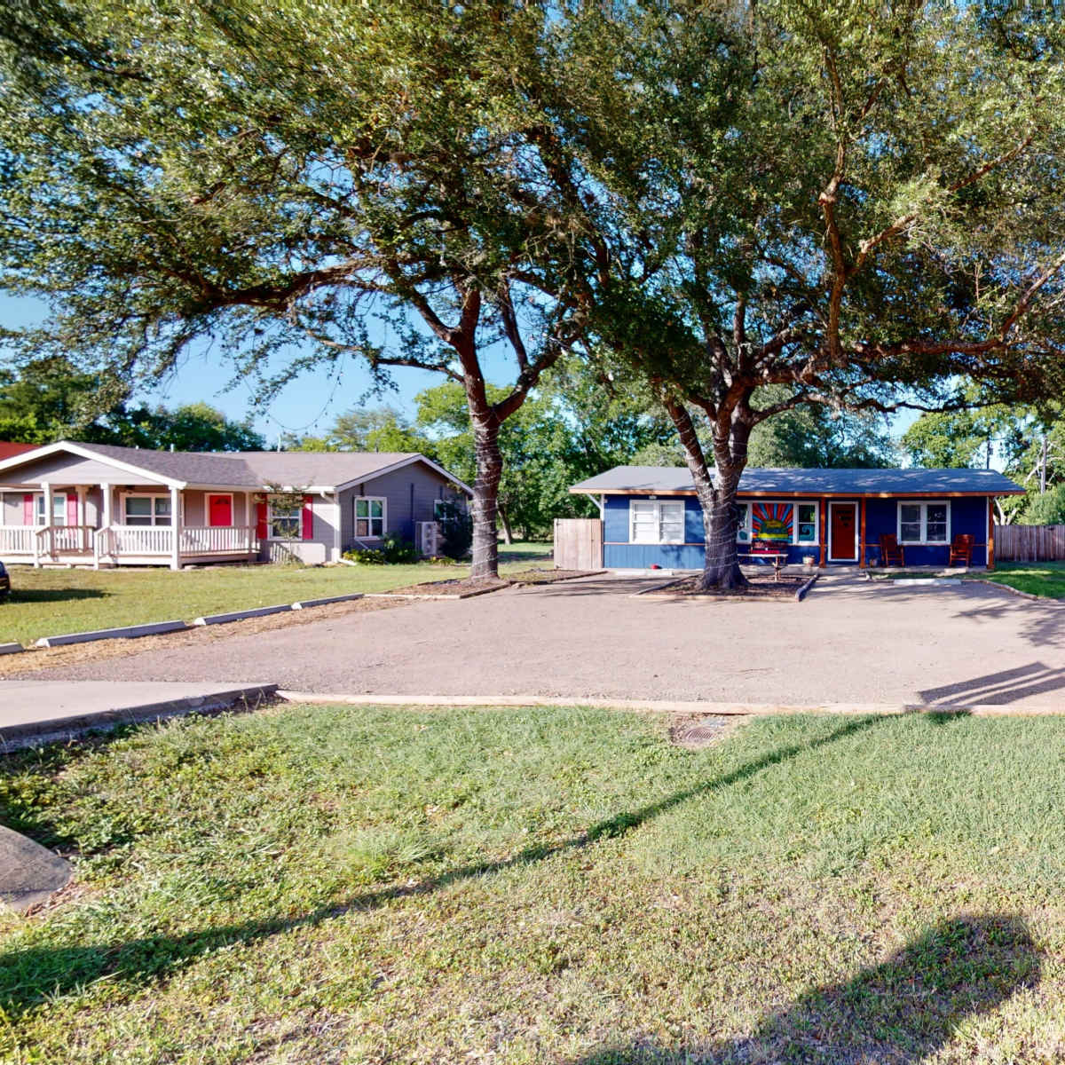 A small commercial building in the center of a gravel lot, flanked by two residential houses and shaded by large trees.