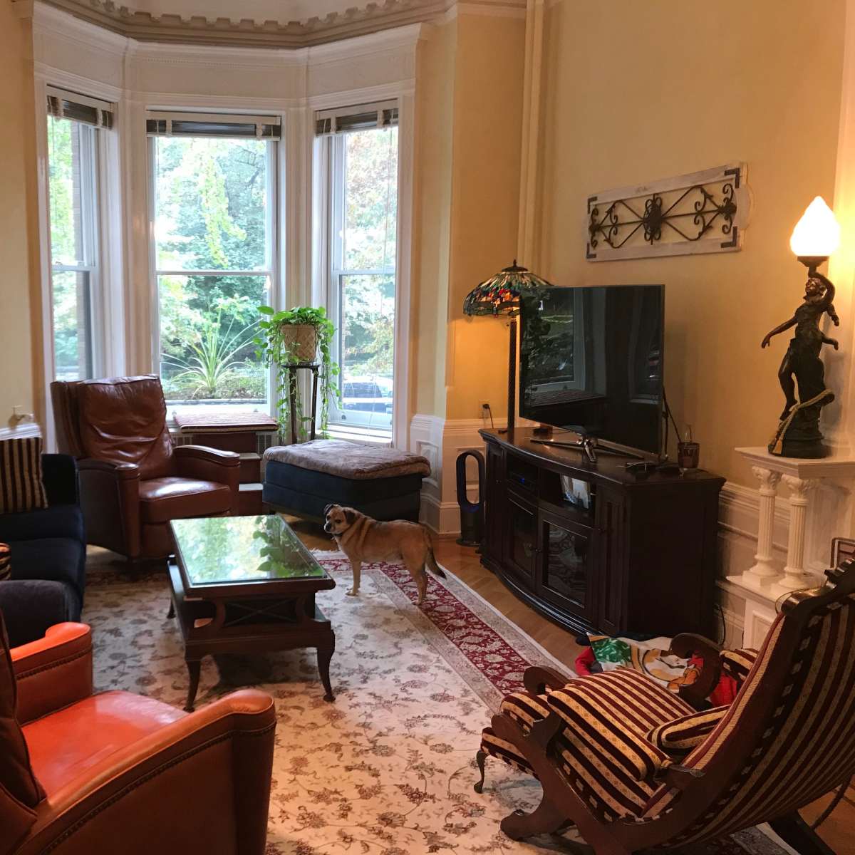 The living room features a mix of brown leather and upholstered armchairs, a green coffee table, and a television near a window overlooking a garden.