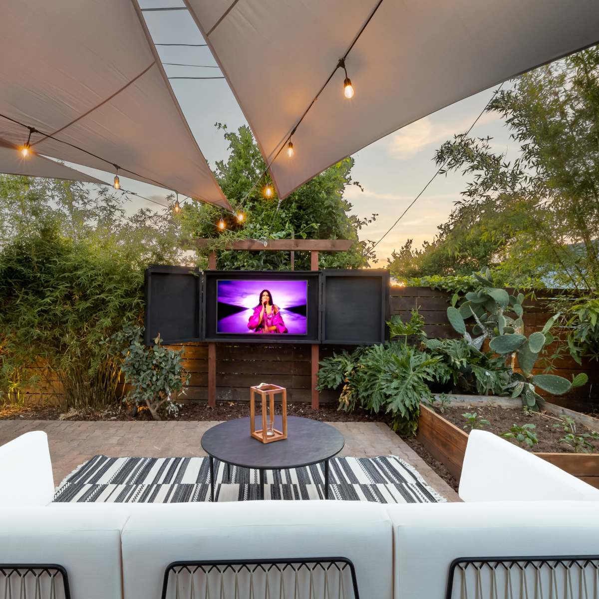 A cozy outdoor patio features a white sofa, a round table, and a television, all under a shaded canopy adorned with string lights and surrounded by greenery.