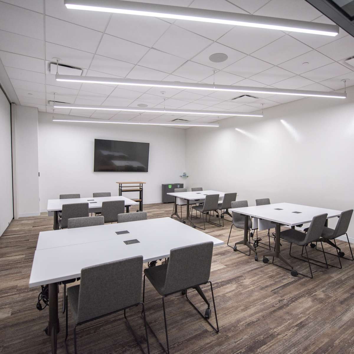A modern meeting room features multiple tables and chairs arranged around a central space, with a wall-mounted screen and soft lighting.