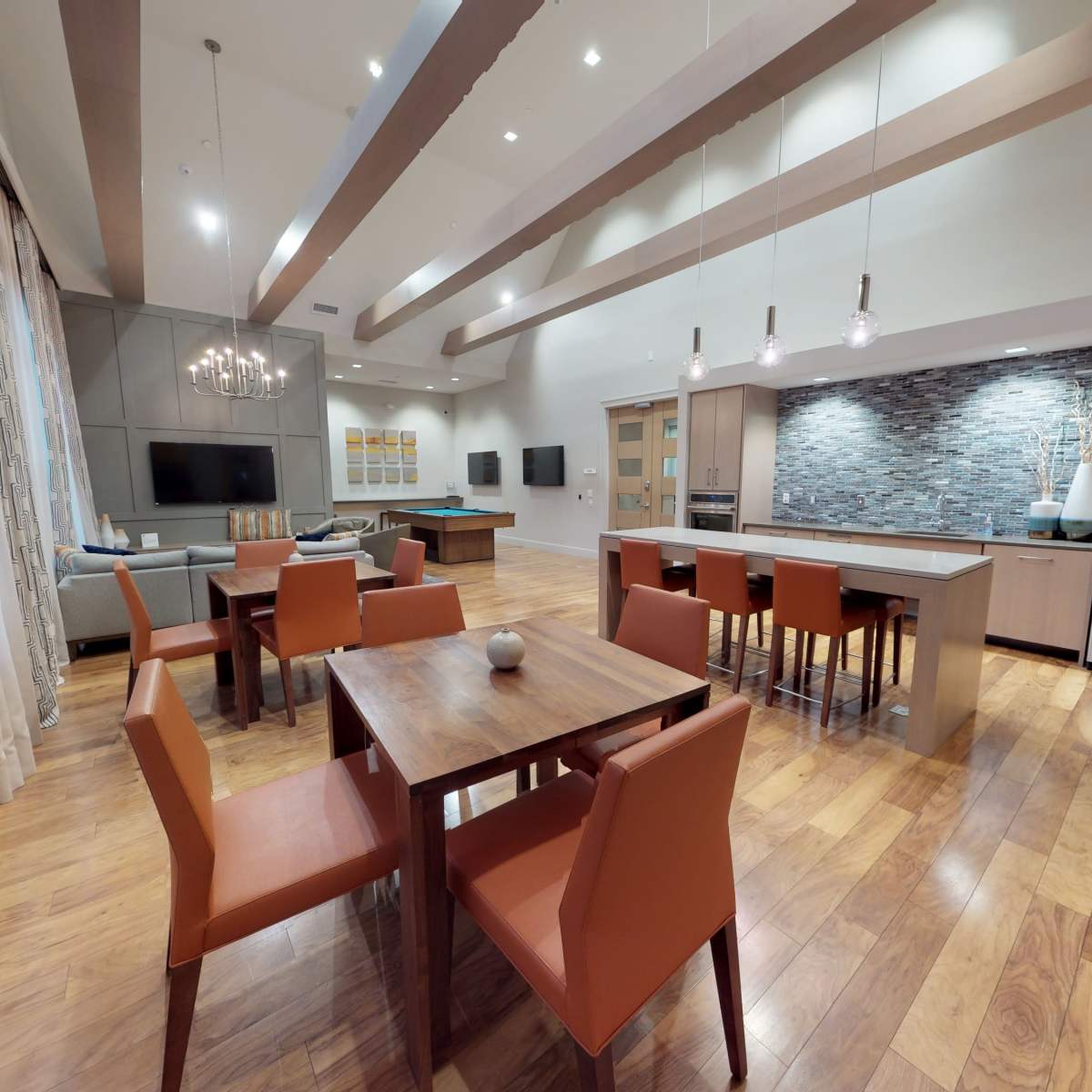 A spacious, modern kitchen and dining area with a wooden dining table surrounded by orange chairs and a kitchen equipped with stainless steel appliances and a stone backsplash.