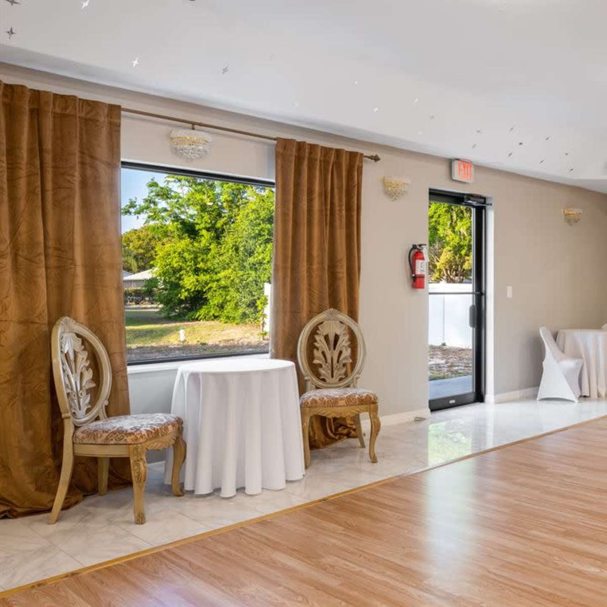The photo shows a spacious room with wooden floors, two ornate chairs near a small table, and large windows covered with brown curtains.