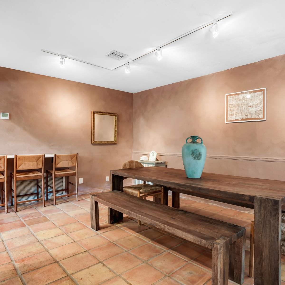 A dining area featuring a long wooden table surrounded by chairs, with bar seating against the wall and a decorative vase on the table.