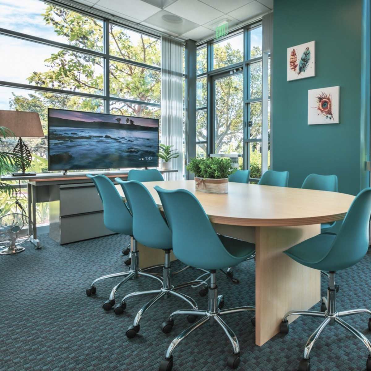 A meeting room features a round wooden table surrounded by six teal chairs, with a large window overlooking greenery and a television displaying a landscape.