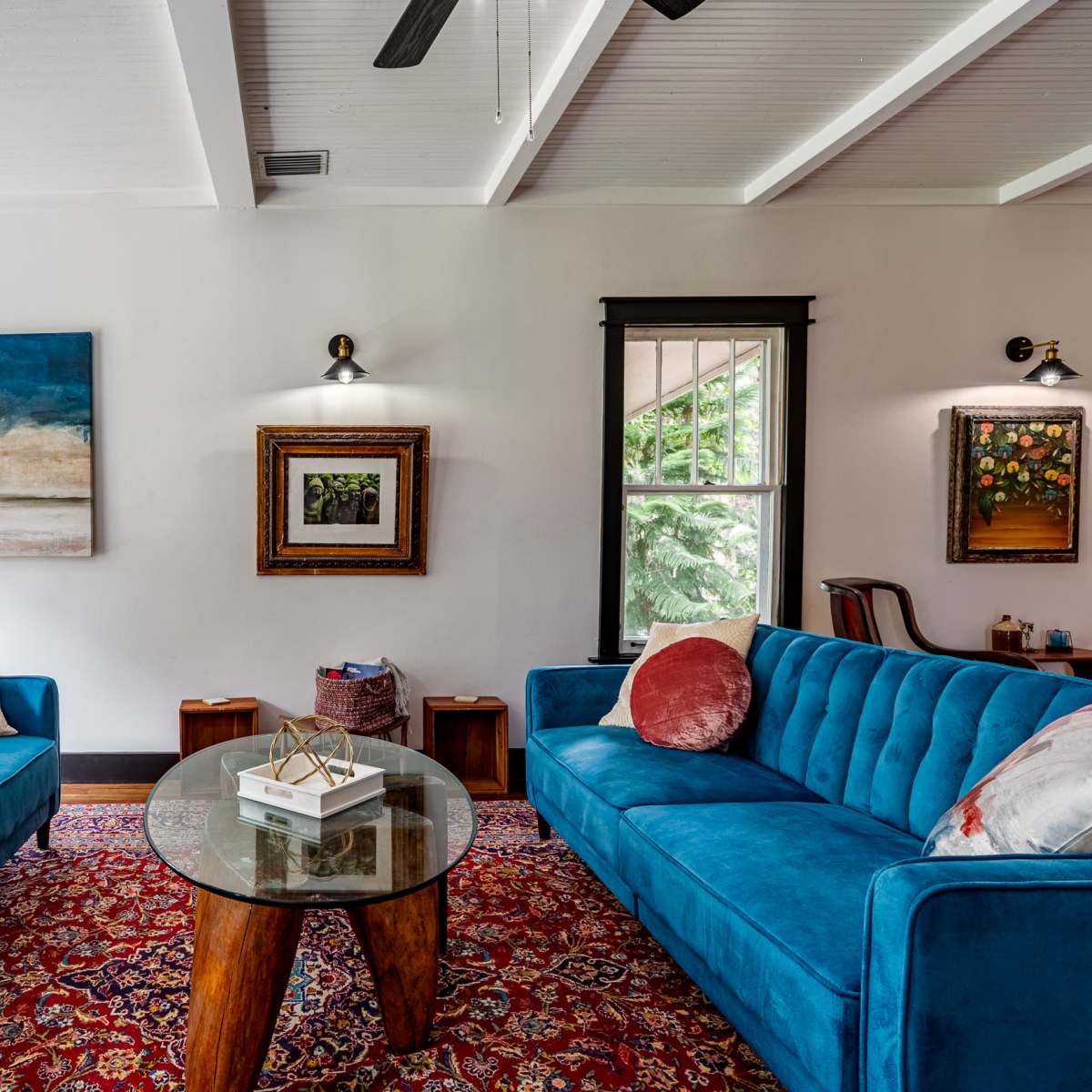 A living room features two blue sofas facing a round glass table on a patterned rug, with artwork and windows allowing natural light.