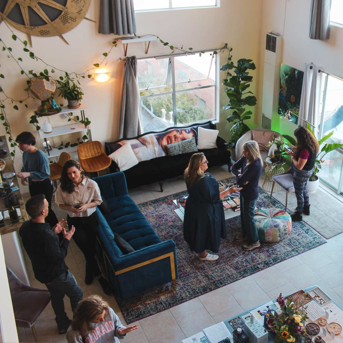A group of people engages in conversation and activities in a spacious, brightly lit living room adorned with plants and colorful decor.