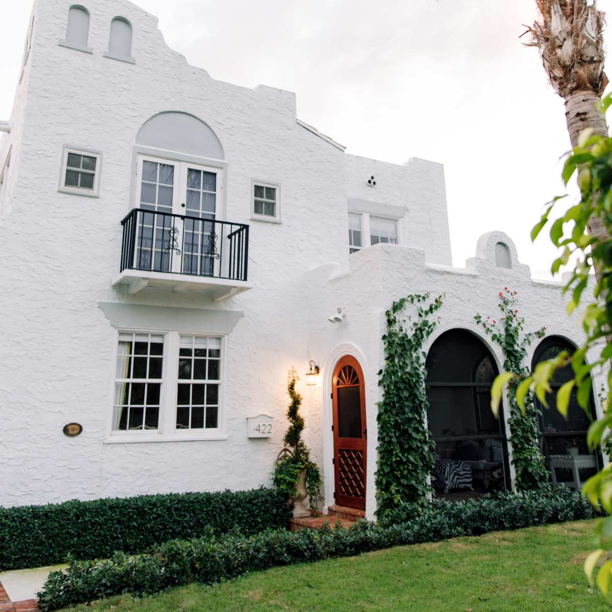 A white, textured house with a black balcony and arched entrances, surrounded by lush greenery and a palm tree.