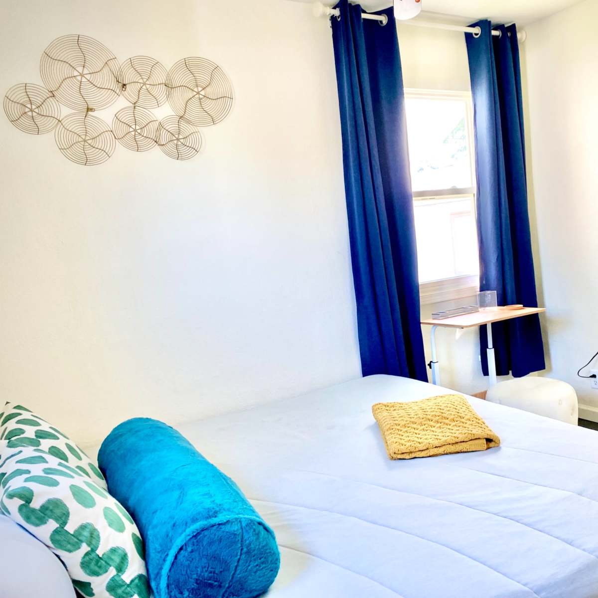 A cozy bedroom with a bed covered in light bedding, a small side table, a TV mounted on the wall, and blue curtains draped over a window.