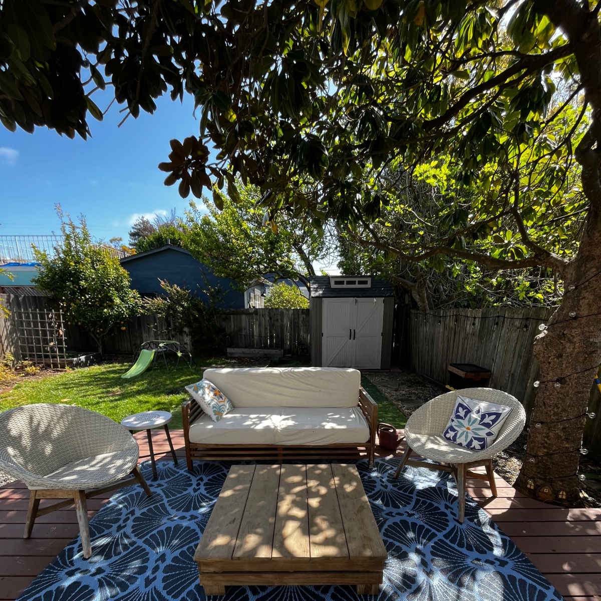 A patio area features a low wooden coffee table, two chairs, and a couch arranged on a patterned rug under a tree.