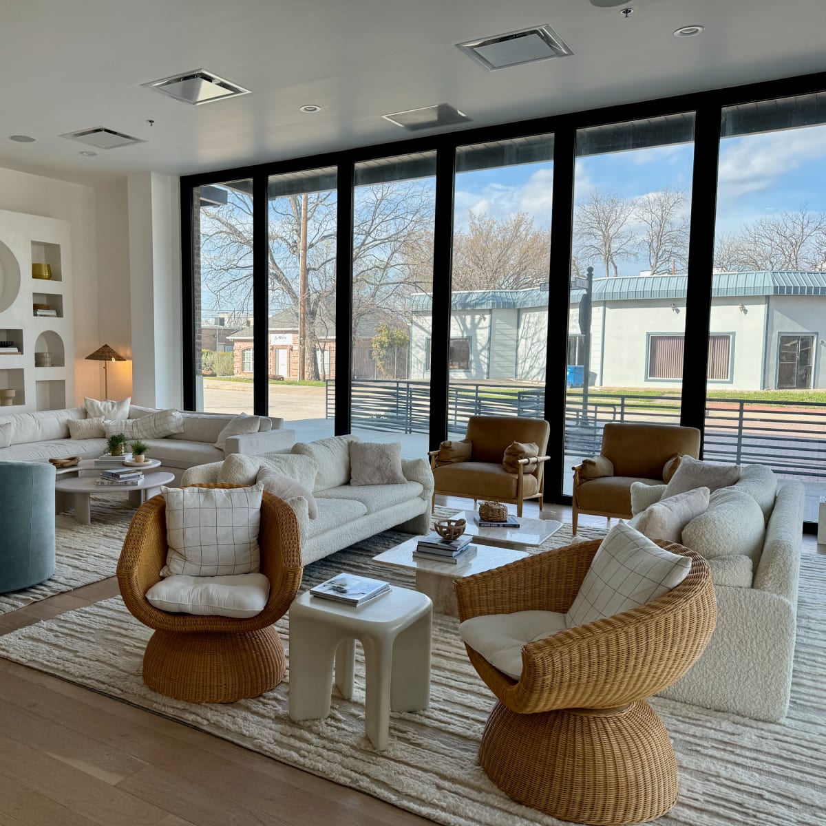 A modern living room features a variety of seating options arranged around a large rug and illuminated by natural light from large windows.