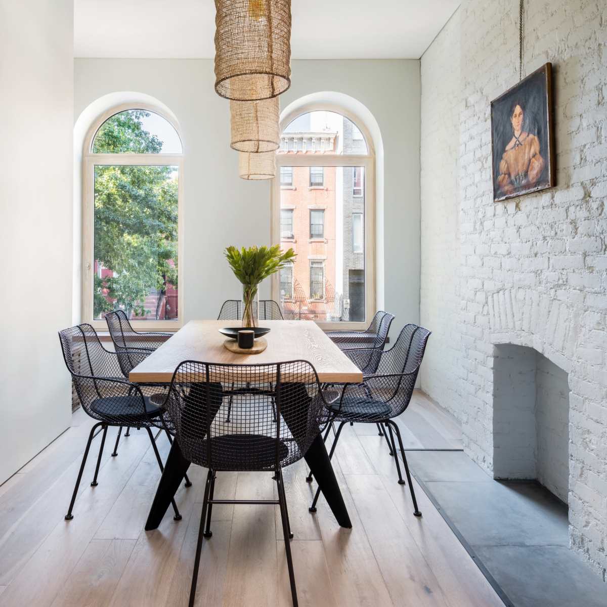 A dining area with a wooden table surrounded by black wire chairs, large windows revealing a view of a street, and three woven pendant lights hanging from the ceiling.