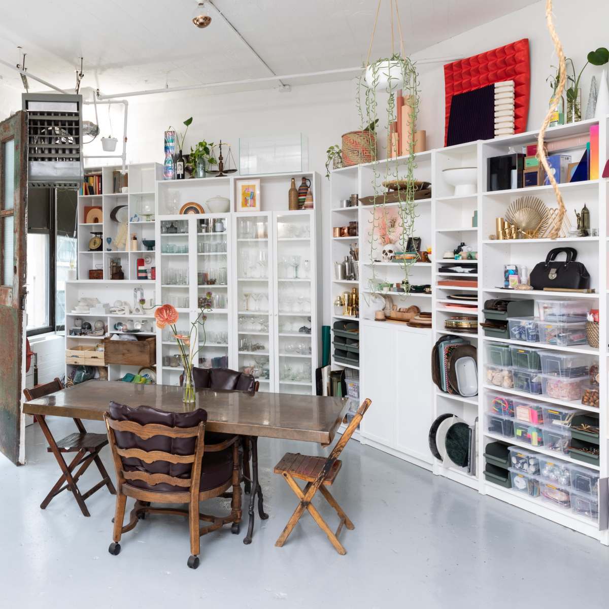 A spacious room with large windows, a wooden table surrounded by chairs, and a shelving unit filled with various items.