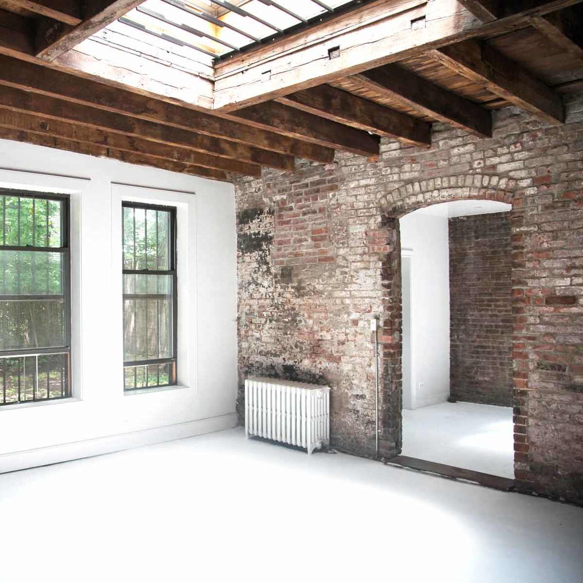 An empty room with large windows, exposed brick walls, wooden beams, and a skylight.