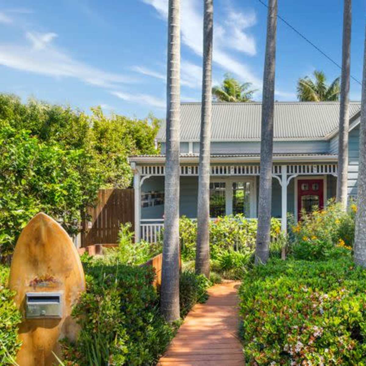 A blue wooden house with a gray roof is surrounded by palm trees and lush greenery, featuring a wooden pathway leading to the front door.