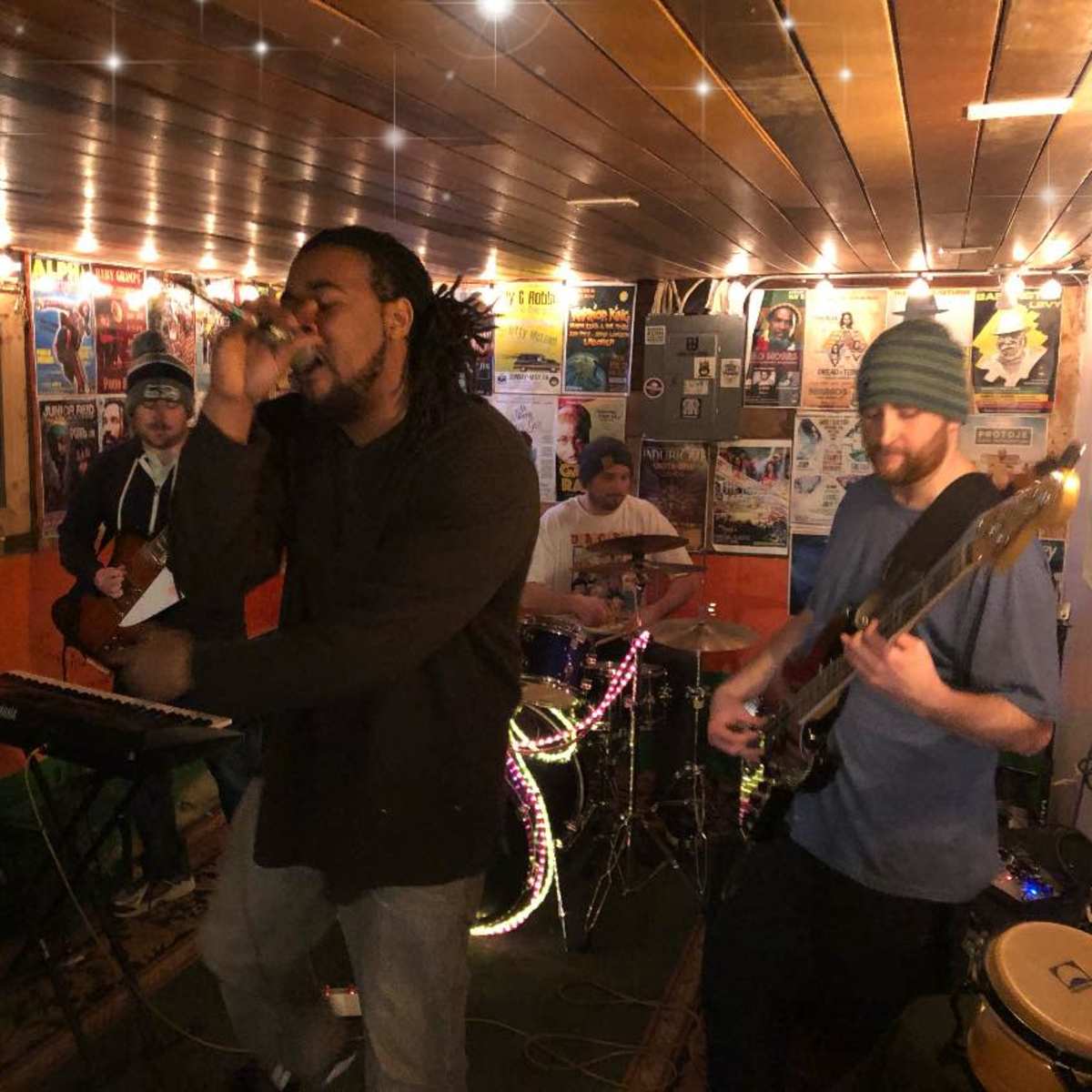 A musical group performs on stage in an intimate venue with instruments and sound equipment visible around them.