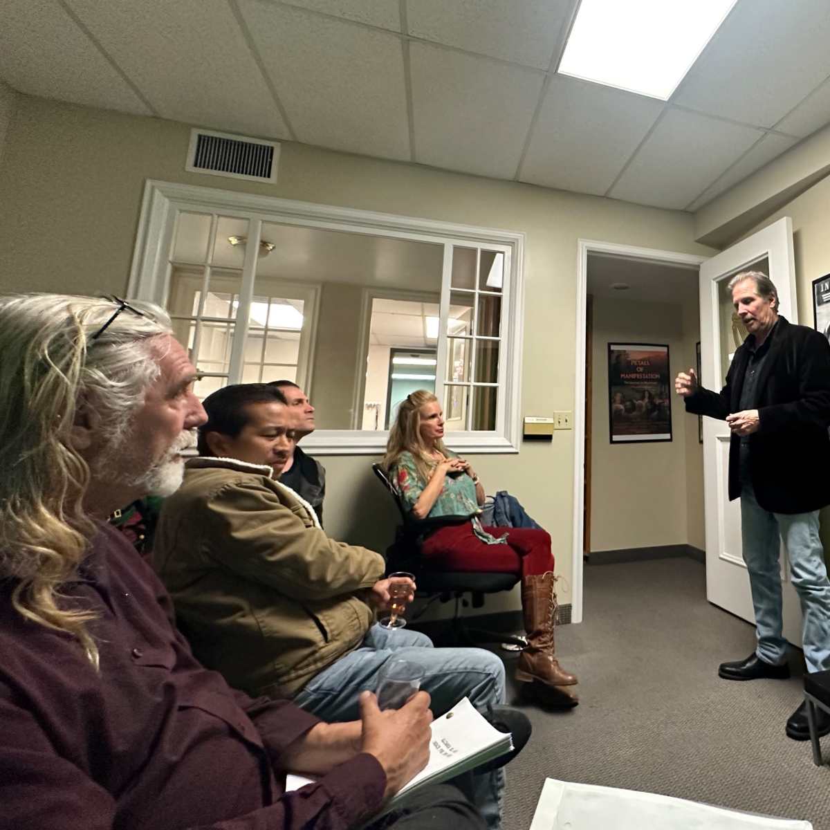 A man stands in front of a screen displaying "THERE IS NO BOX STUDIOS," while a small audience of three sits attentively in a room with light-colored walls and framed posters.