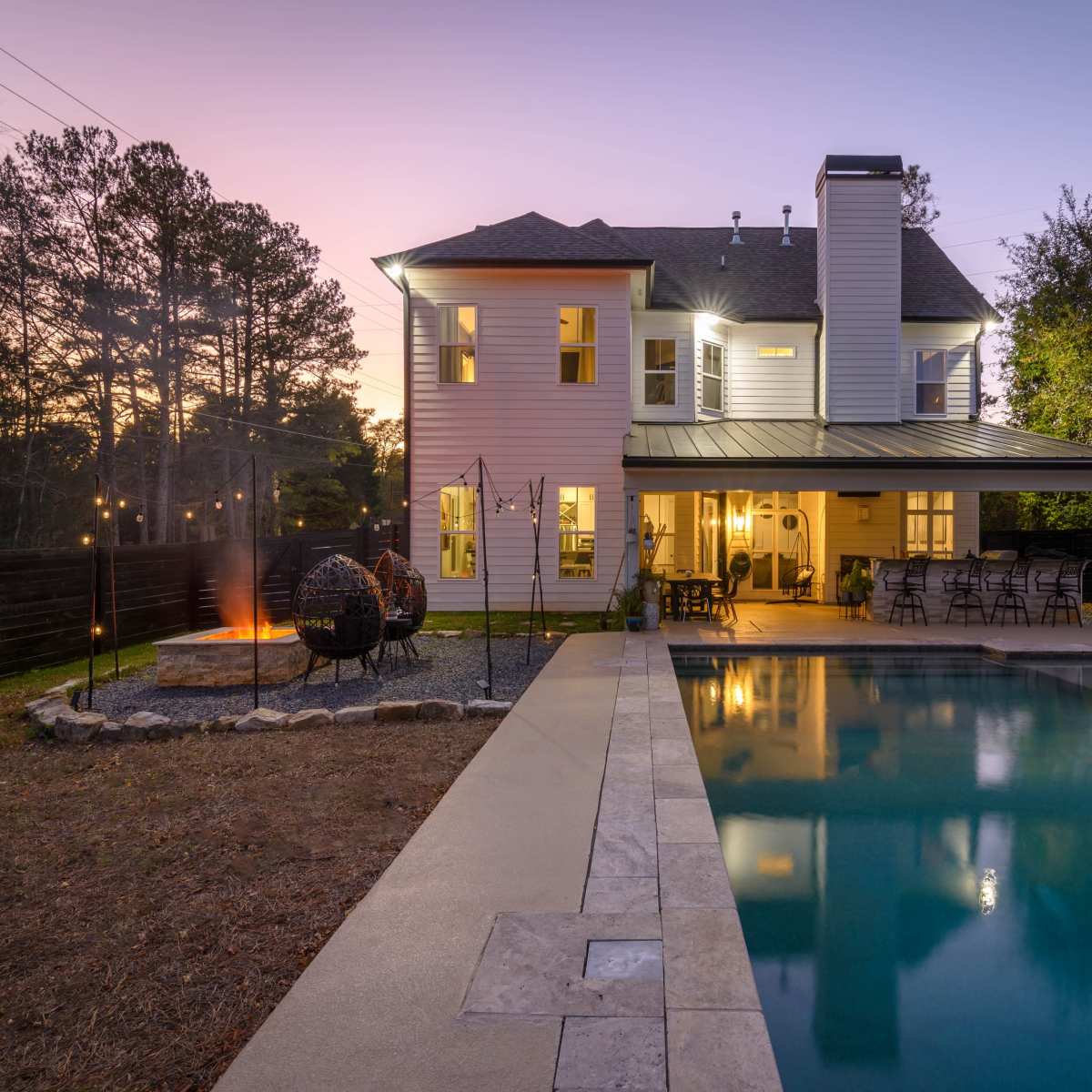 A modern two-story house with a pool and outdoor seating area, surrounded by trees at dusk.