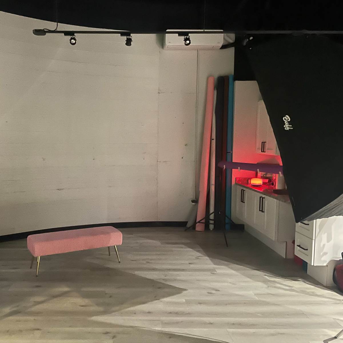 A sparsely decorated photography studio featuring a pink bench, large softbox lights, and a kitchen area in the background.