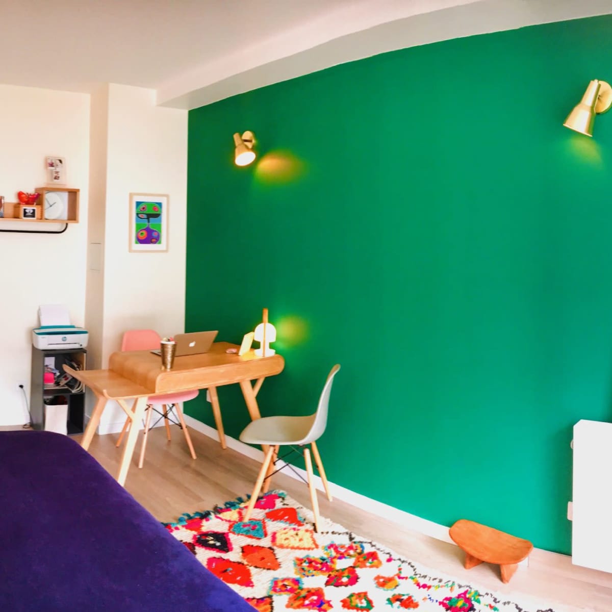 A small room featuring a green accent wall, a wooden table with a laptop, a bookshelf, and colorful decor on the floor.