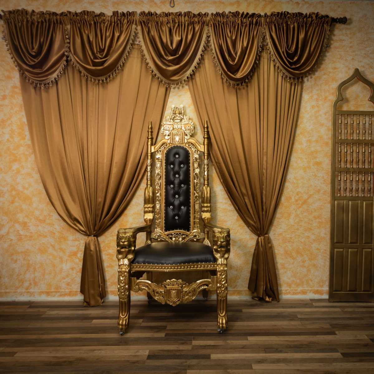 A gilded throne is positioned in front of elaborately draped golden curtains against a textured orange wall, complemented by decorative wooden furniture.