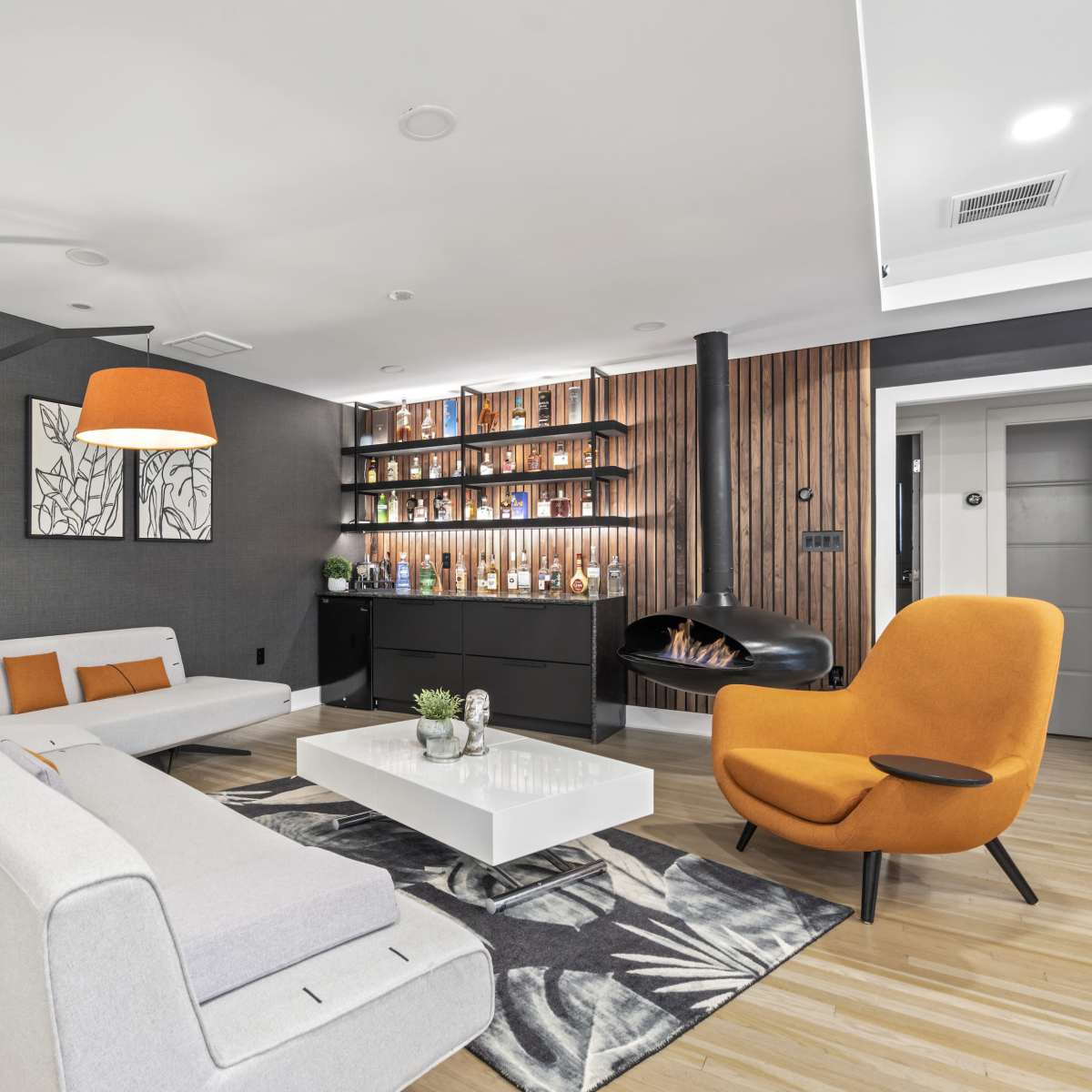 A modern living room features a gray wall, a white sofa, an orange chair, a coffee table, and a display of decorative bottles on open shelves.