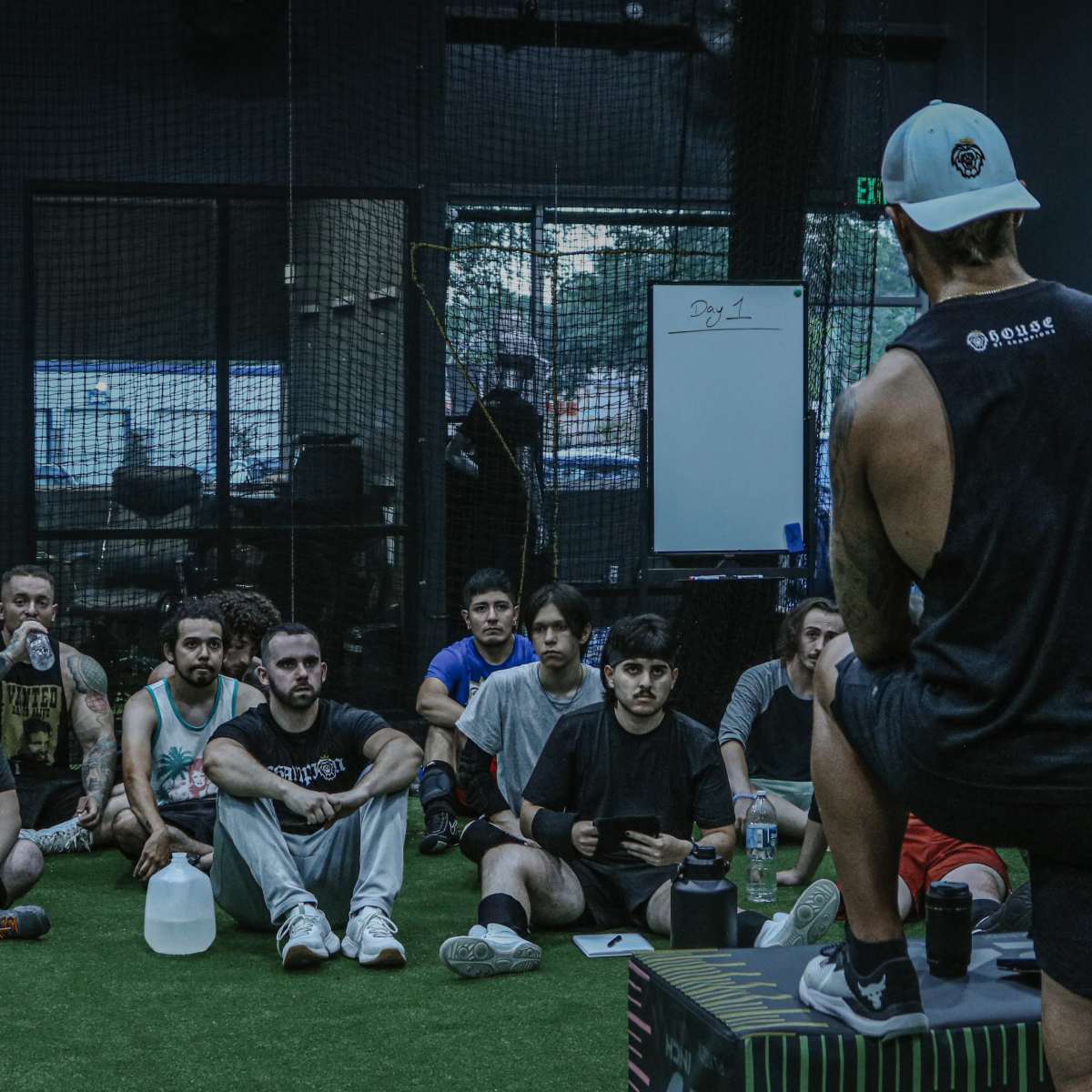 A fitness coach stands on a platform addressing a group of seated athletes in a gym.