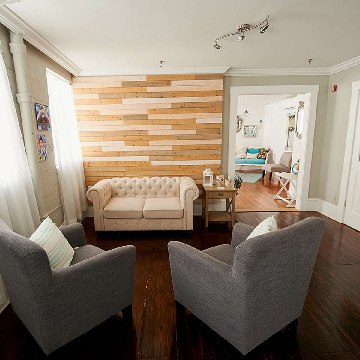 A living room features a chesterfield sofa and two gray armchairs, with a wooden accent wall and large windows letting in natural light.
